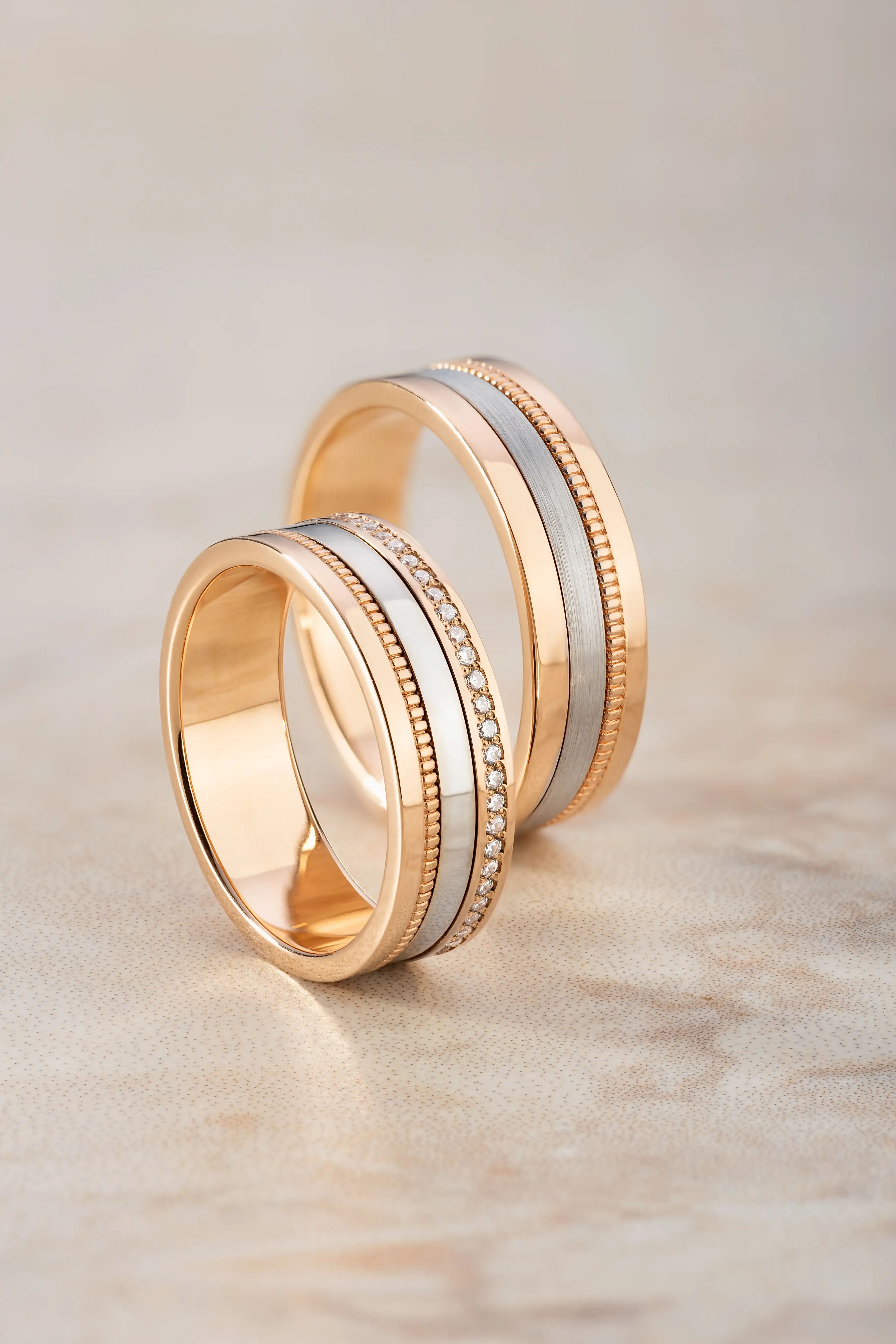 Pair of rose gold wedding bands with silver inlays; one band features a row of small diamonds, both displayed on a soft beige textured surface.